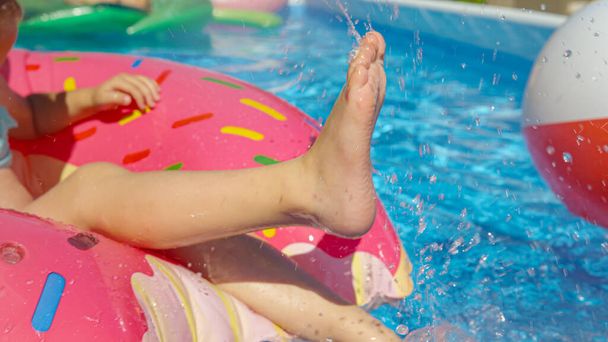 CLOSE UP: маленькі дівочі ноги розбризкують воду з пончика плаваті в басейні на задньому дворі. Маленька дівчинка робить бризки води, розважаючись на вечірці в басейні. Літні розважальні заходи для спекотних днів в саду
 - Фото, зображення
