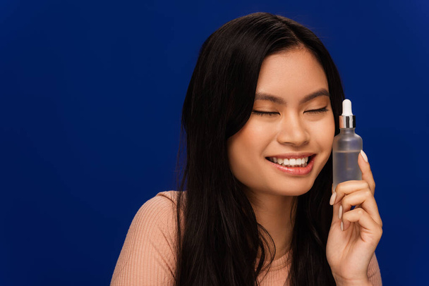 Весела азіатська жінка тримає пляшку з косметичною сироваткою ізольована на синьому
  - Фото, зображення