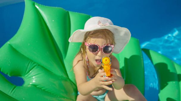 TOP DOWN Little girl sitting on a floatie with water gun pointing towards camera. Cheerful little girl floating in backyard swimming pool and having fun with water blaster spraying in camera direction - Photo, Image