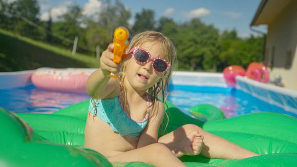 CLOSE UP: Adorable girl sitting on a floatie in garden pool holding a water gun. Playful little girl floating in backyard swimming pool and having fun with water blaster spraying directly into camera - Foto, Bild