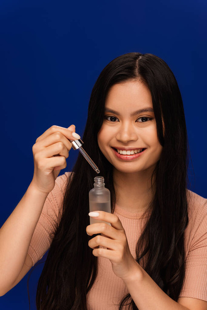 Smiling asian woman holding cosmetic serum and dropper isolated on blue  - 写真・画像