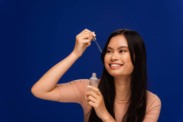 Веселая азиатка, держащая косметическую сыворотку и глядя на пипетку, изолированную на голубом  - Фото, изображение