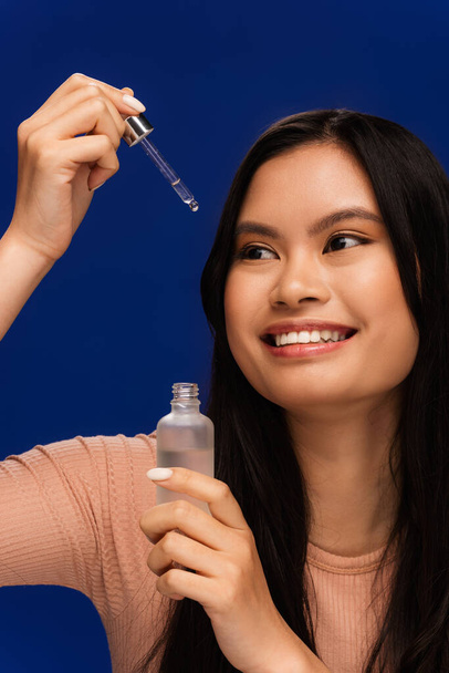 Pretty asian woman in t-shirt holding dropper and cosmetic serum isolated on blue  - Foto, Bild