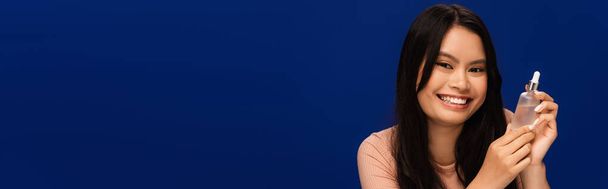 Smiling brunette asian woman holding cosmetic serum isolated on blue, banner  - Fotografie, Obrázek