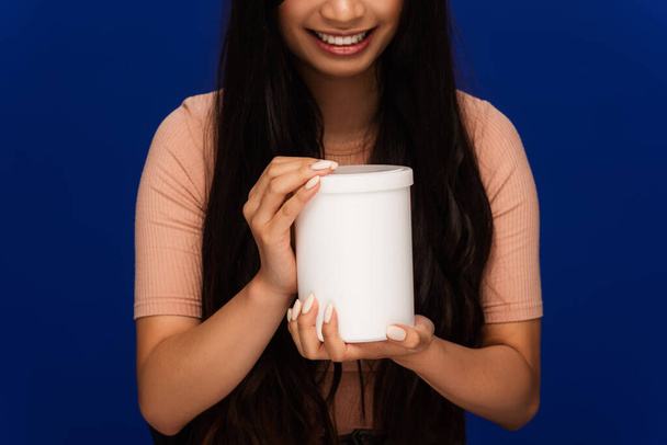 Μερική άποψη της θετικής γυναίκας που κατέχουν βάζο του καλλυντικού προϊόντος που απομονώνονται σε μπλε  - Φωτογραφία, εικόνα