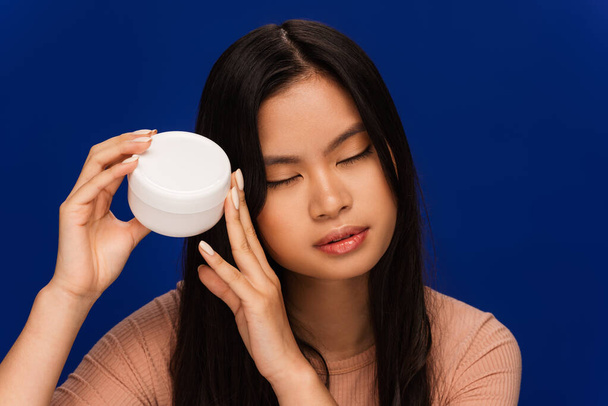Pretty asian woman holding container with cosmetic cream isolated on blue  - Fotoğraf, Görsel