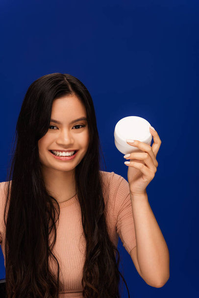 Cheerful long haired asian woman holding container with cream isolated on blue  - Foto, Imagem