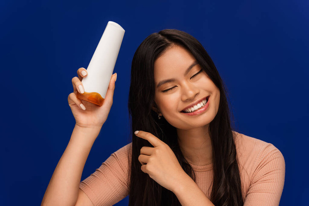 Smiling asian woman holding hair conditioner isolated on blue  - Foto, afbeelding