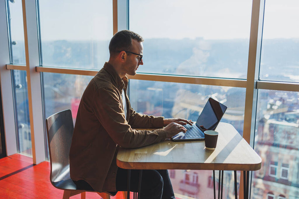 A balanced man in glasses and casual clothes works on a laptop from a cozy workspace. A successful freelancer works remotely. - Fotoğraf, Görsel