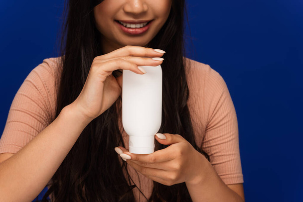Cropped view of positive brunette woman holding cosmetic lotion isolated on blue  - Photo, image