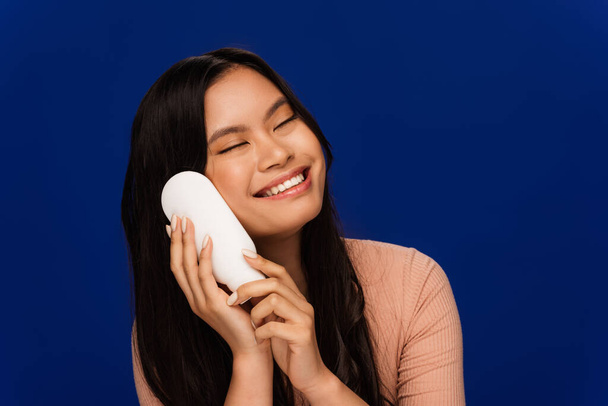 Cheerful brunette asian woman holding body lotion isolated on blue  - Fotoğraf, Görsel