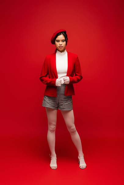 Full length of stylish asian model in beret looking at camera on red background  - Photo, image