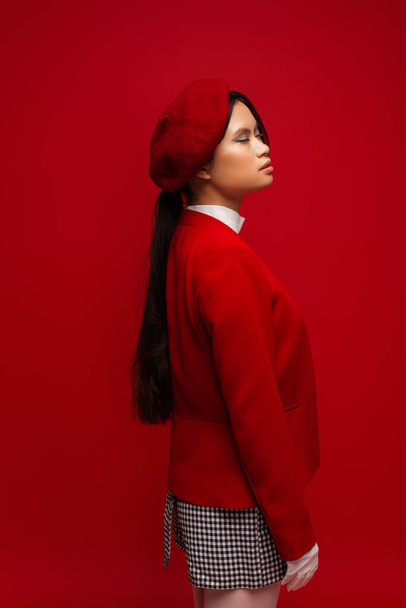Brunette asian model in jacket and beret closing eyes isolated on red  - Foto, immagini