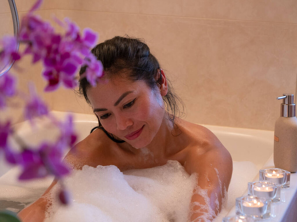 Attractive young lady washing her body in bath full of bubbly foam. Beautiful Philippine lady body washing herself with bath foam. Time for personal hygiene in relaxing home ambience. - Φωτογραφία, εικόνα