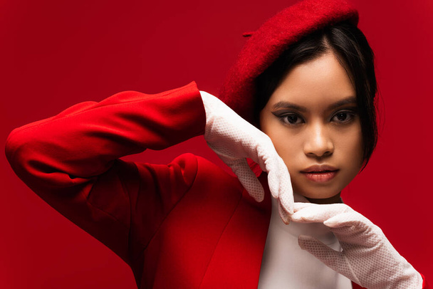 Portrait of asian model in beret and gloves holding hands near face isolated on red  - Photo, Image