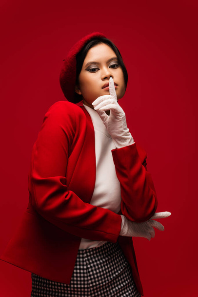 Stylish asian model in beret and jacket showing shh gesture isolated on red  - Valokuva, kuva