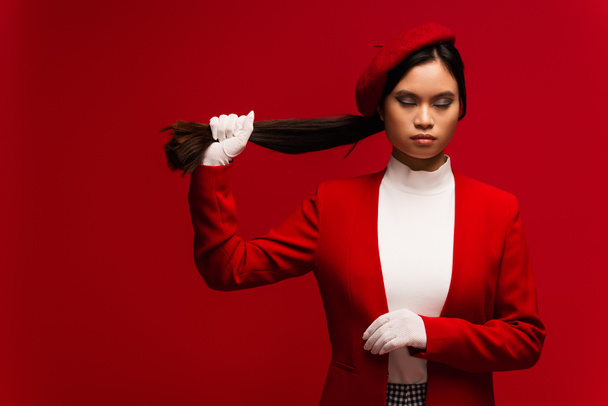 Asian model in gloves and beret touching hair isolated on red  - Foto, Bild