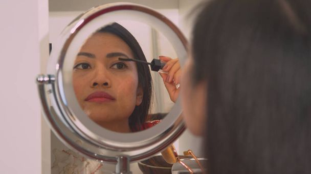 Beautiful Philippine woman in mirror reflection applying black mascara. Young lady putting on make up for an evening date in front of a mirror. Female person taking care of her appearance. - Fotografie, Obrázek