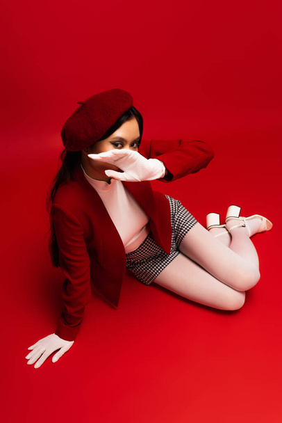 overhead view of trendy asian model in white coves covering face while sitting on red background  - Fotó, kép