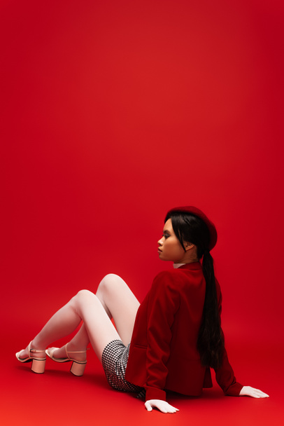Side view of stylish asian model in white tights and gloves looking away while sitting on red background  - Foto, Bild
