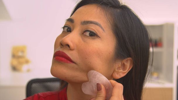 Young lady massaging her face with gua sha scraper for facial lifting. Beautiful Asian woman using gua sha scraper for rejuvenating anti-aging massage and facial muscles relief treatment. - Foto, Imagen