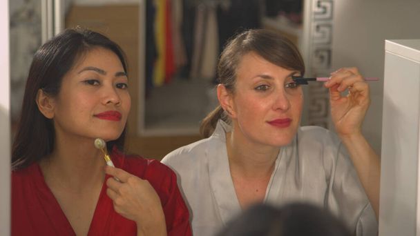CLOSE UP Detailansicht schöner Frauen, die sich in Spiegelreflexion schminken. Kaukasische und asiatische Frauen verwenden Schönheitsprodukte und bereiten sich auf einen Abend vor. Zwei Damen bei einer Schönheitsbehandlung - Foto, Bild