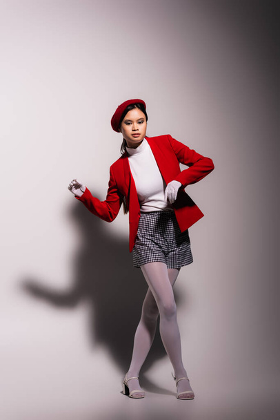 Trendy asian model in white gloves and red jacket posing on grey background with shadow  - Photo, Image