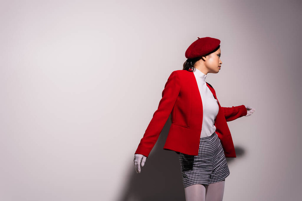 Side view of fashionable asian woman in beret and shorts standing on grey background with shadow  - Fotografie, Obrázek