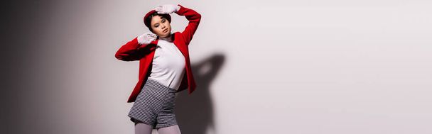Fashionable asian woman in jacket and beret standing near shadow on grey background, banner  - Foto, Bild