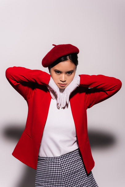 Портрет модной азиатской модели в красно-белой одежде, смотрящей в камеру на сером фоне  - Фото, изображение