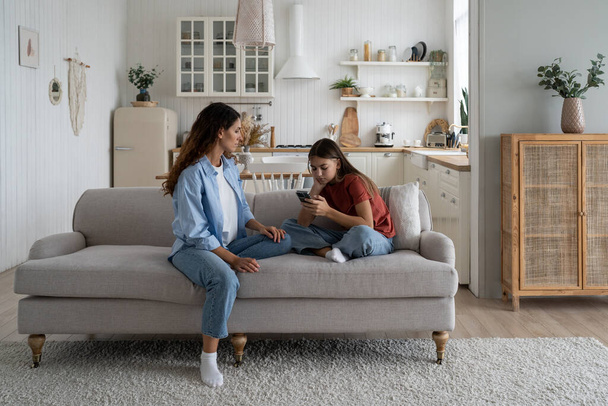 Sad teen girl holding mobile phone, experiencing cyberbullying on social media, sitting with mother on sofa at home. Mom helping daughter to cope with unrequited love. Teenage problems concept - Fotografie, Obrázek