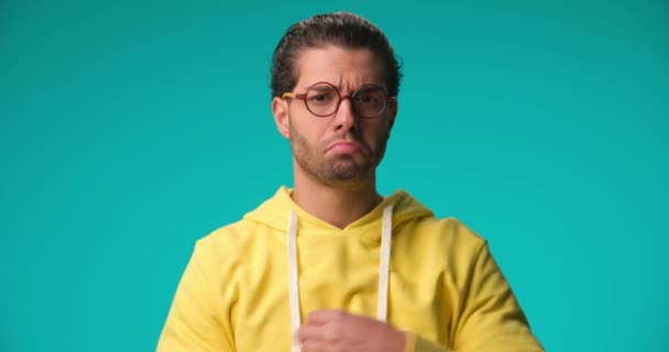 powerful guy in yellow hoodie showing his muscles and strength, nodding and folding arms on blue background - Imágenes, Vídeo