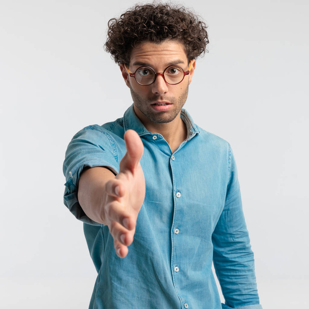 Portrait of handsome casual man raising his arm to shake hands, standing, wearing a shirt and eyeglasses against gray studio background - Foto, afbeelding