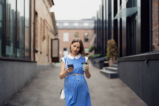 Ocupada mujer embarazada moderna caminando entre edificios de oficinas usando un teléfono inteligente. Mujer en un vestido vintage azul y monedero blanco usando el teléfono sobre la marcha - Foto, Imagen