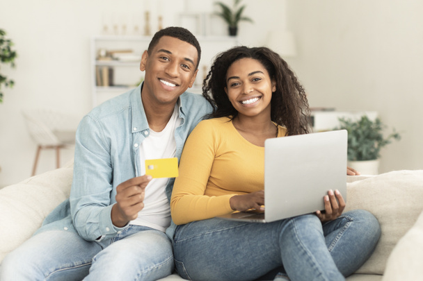 Cheerful young african american family use credit card, laptop and banking, check financial account, sit on sofa in living room interior. Sale for shopaholics, online shopping, ad and offer at home - Zdjęcie, obraz