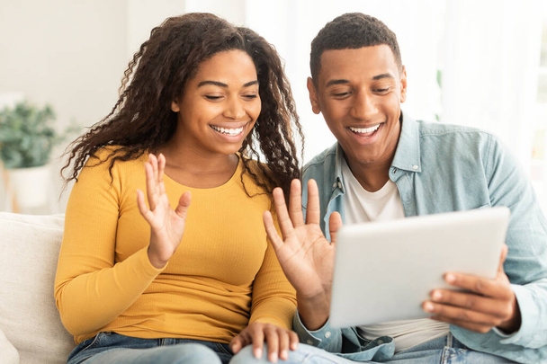 Cheerful young african american guy and lady waving hands at tablet webcam, greeting, say hello on sofa in living room interior, close up. Application for communication and meeting, new normal at home - Zdjęcie, obraz