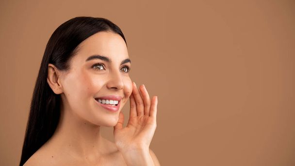 Headshot of cheerful beautiful brunette half-naked millennial woman touching her smooth young skin, looking at copy space and smiling, enjoying spa treatment results, beige studio background - Photo, image