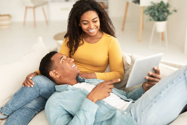 Glad young african american guy shows tablet to lady, recommends website and app, rest and relax together in free time on sofa in room interior. Video in social networks, surfing in internet at home - Foto, Imagen