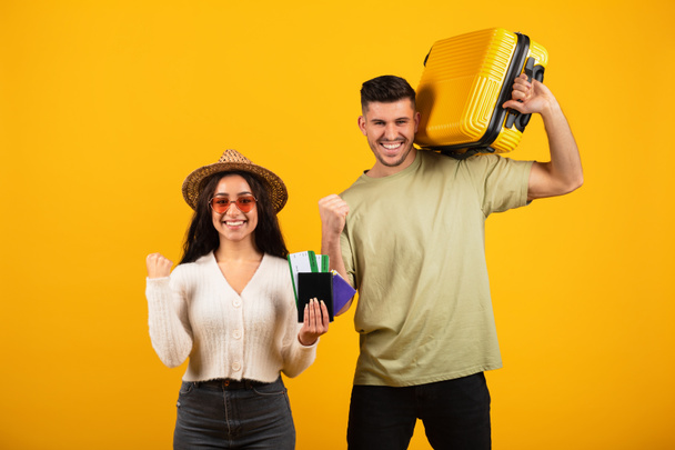 Smiling millennial arab woman in hat, glasses show passports, tickets, man with suitcase making victory hand gesture, isolated on orange background, studio. Sign of success, joy of journey in vacation - Fotó, kép