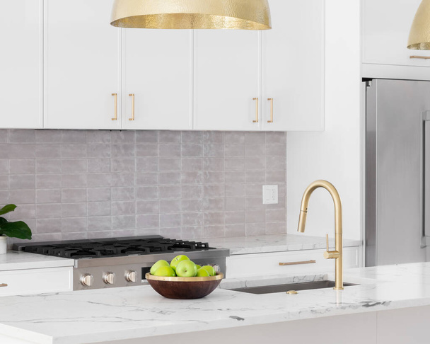 A kitchen detail with white cabinets, gold faucet and light hanging over the island, and a tiled backsplash. - Foto, afbeelding