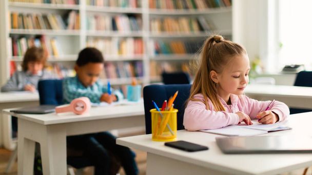 Разнообразные дети сидят за столами в классе в начальной школе или детском саду, пишут или рисуют в блокнотах. Восстановление и возвращение в школу после блокировки - Фото, изображение