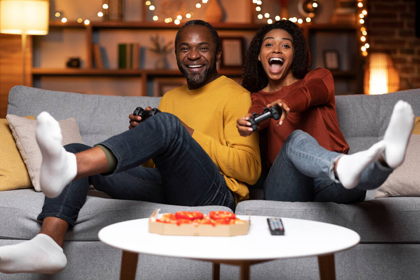 Emotional happy black spouses sitting on couch, holding joysticks and laughong, couple playing video games at home, eating pizza, living room decorated with festive lights, copy space - Photo, image
