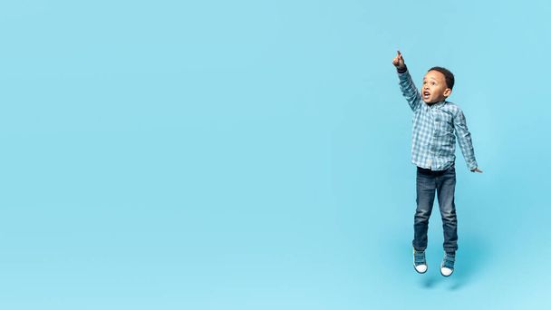 Emotional little african american kid boy jumping up and pointing at copy space for advertisement over blue studio background, showing something exciting, panorama, full length - Photo, image