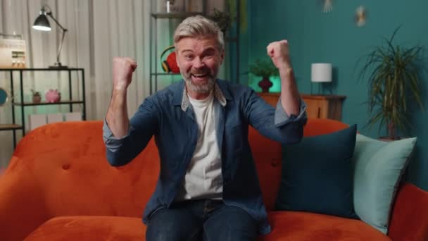 Happy senior man shouting, celebrating success, winning, goal achievement, good news raising fists in gesture I did it, lottery luck at home apartment. Mature middle-aged guy in night room on couch - Filmmaterial, Video