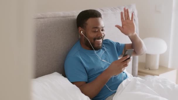 Distant communication. Young happy african american guy in earphones video chatting with friend on smartphone, resting in bed in morning, tracking shot, slow motion, free space - Video, Çekim