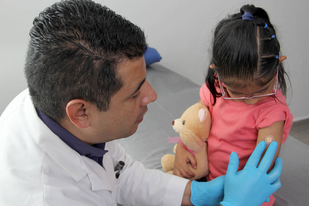 黒い髪のラテン系の医師と少女は、小児科の診察を受けて、コヴィド、水痘、ジフテリア、インフルエンザ、肝炎、はしか、塊に対して腕を予防接種する - 写真・画像