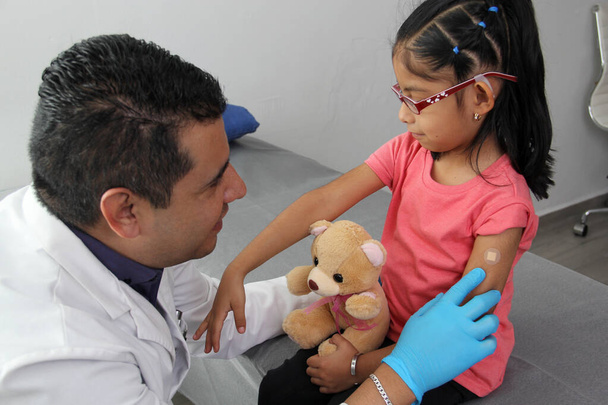 Dark-haired Latino doctor and little girl have a medical consultation in the pediatric office to vaccinate their arm against Covid, chickenpox, diphtheria, influenza, hepatitis, measles, mumps - Fotó, kép