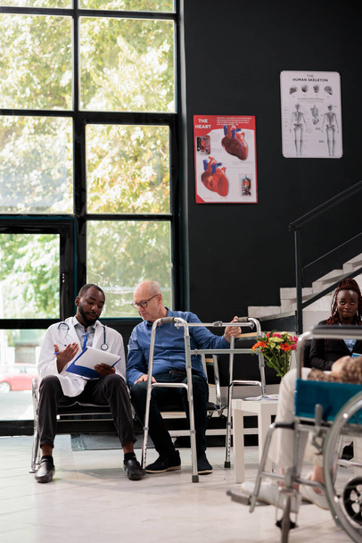African american doctor holding clipboard explaining medical expertise to senior patient with wallking frame during consultation in hospital waiting area. Medicine service and concept - Foto, Imagem