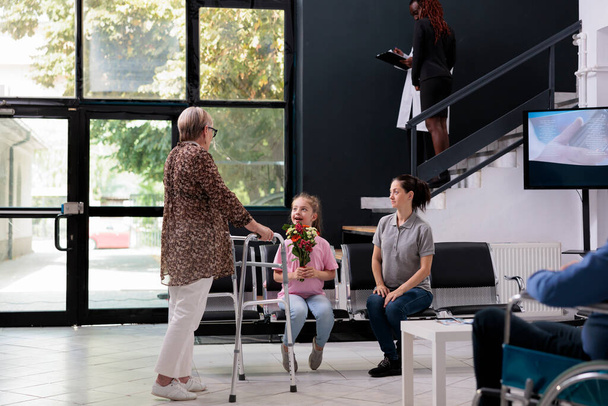 Granddaughter giving flowers to grandmother after finishing medical examination with practitioner doctor. Elderly patient with walking frame discussing disease expertise with family - Foto, afbeelding