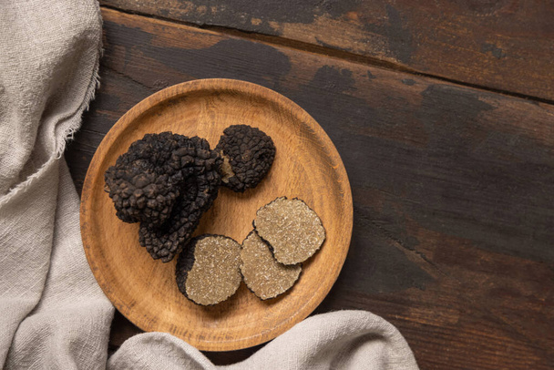 Whole and sliced black truffles mushroom on wooden plate on dark brown table near grey napkin, flat lay, copy space. Culinary delight ingredient.  - Photo, image